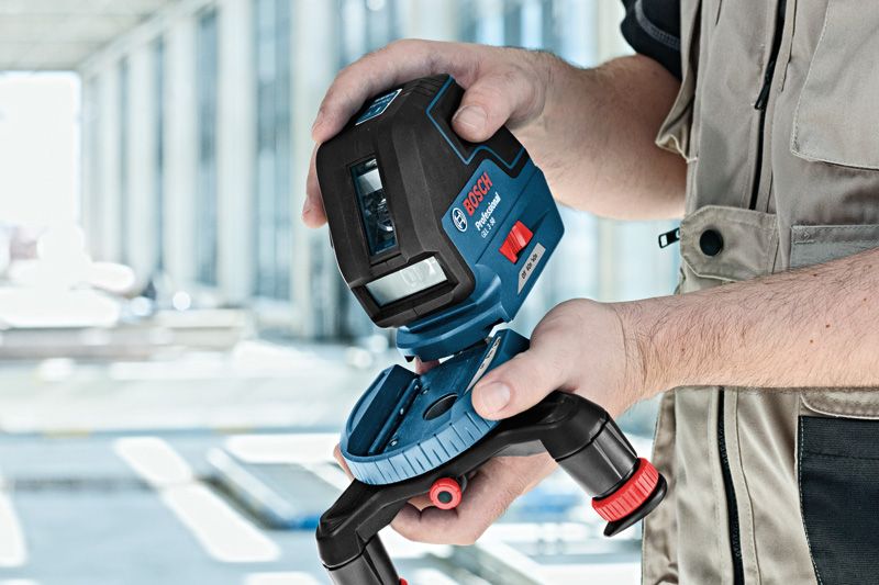 Person in workwear adjusts a laser leveling tool on a tripod indoors.