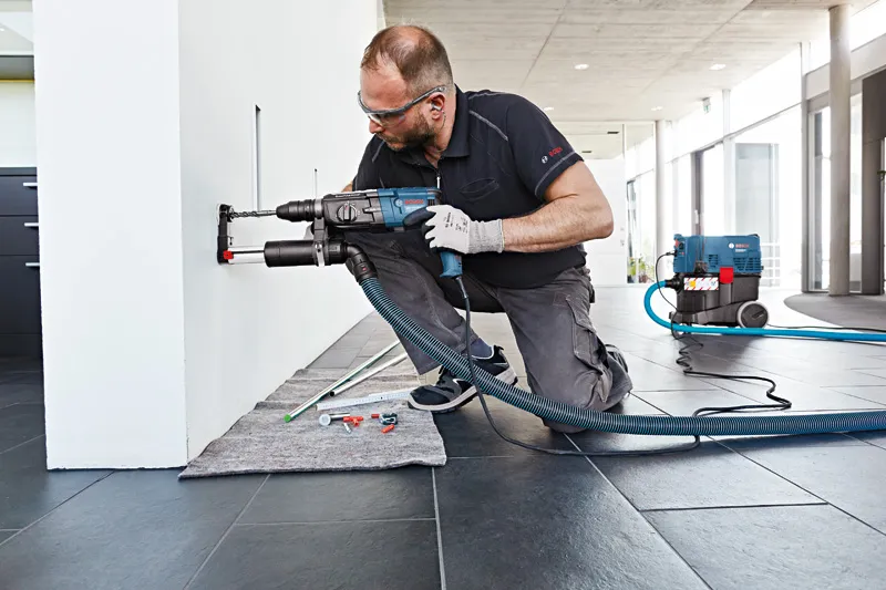 A person wearing safety equipment drills into a wall with a dust extraction attachment.