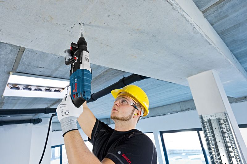 A person wearing safety equipment drills into a concrete ceiling with a rotary hammer.