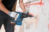 A person wearing safety equipment drills into a concrete wall with a cordless rotary hammer.