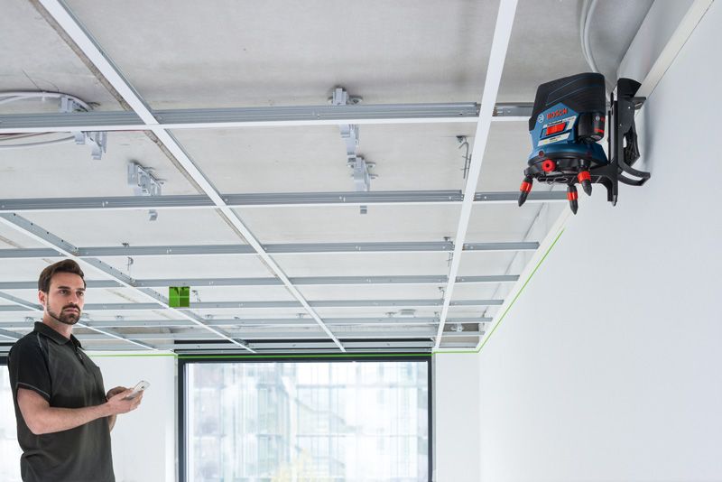 Person using a phone observes a laser leveling tool projecting a green line on a suspended ceiling frame.
