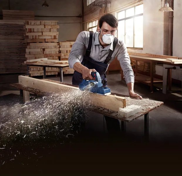 A person wearing safety equipment smooths a wooden plank with a planer.