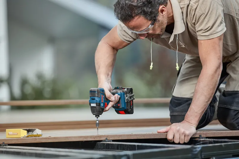 Person wearing safety equipment uses a cordless impact driver to fasten a screw into wood.
