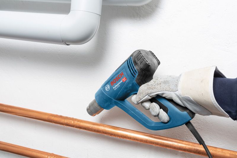 A person wearing safety equipment uses a heat gun on copper pipes.