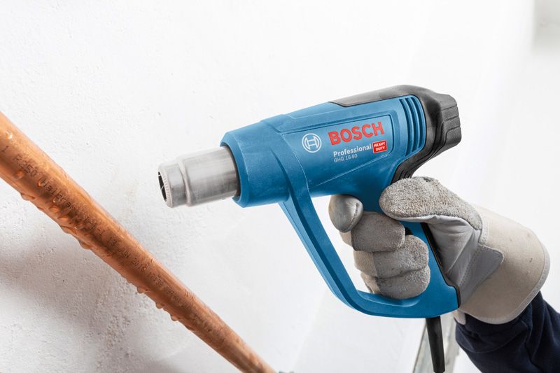 A person wearing safety equipment uses a blue heat gun near a copper pipe.
