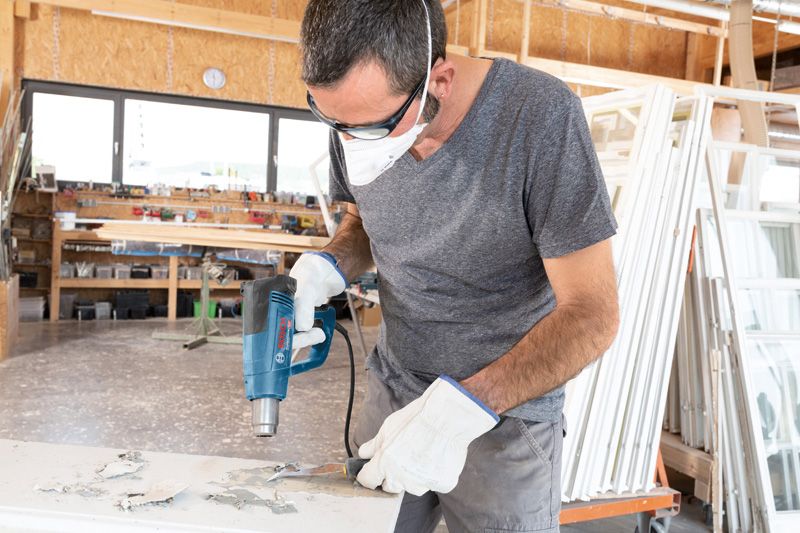 A person wearing safety equipment uses a heat gun to remove paint from a surface.