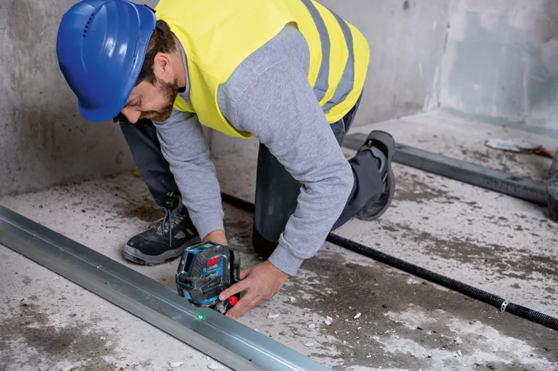 Person wearing safety equipment aligns metal framing with a laser leveling tool on a concrete floor.