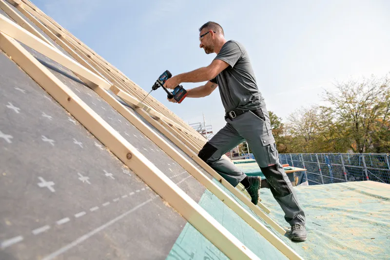 A person wearing safety equipment uses a cordless drill to fasten wood on a sloped roof.