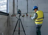A person wearing safety equipment marks a wall using a laser leveling tool on a tripod.