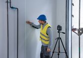 A person wearing safety equipment marks a wall guided by a laser leveling tool on a tripod.
