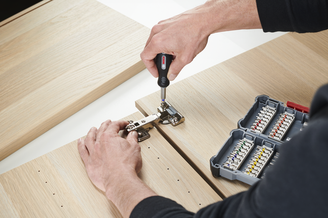 Person assembling a cabinet hinge with a screwdriver next to a set of bits.