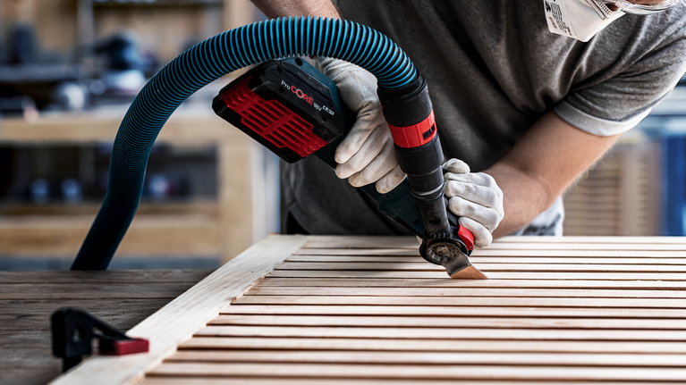 Person wearing safety equipment uses a power tool with vacuum attachment to sand wooden slats.