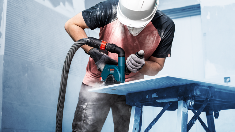 A person wearing safety equipment cuts a sheet with a power tool, dust rising.