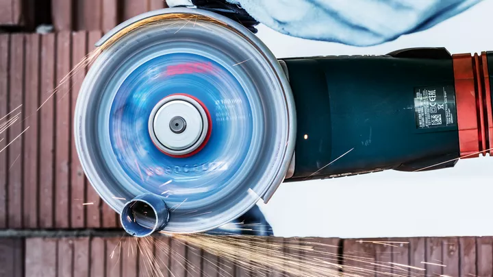 A person wearing safety equipment cuts a metal pipe with a spinning angle grinder.