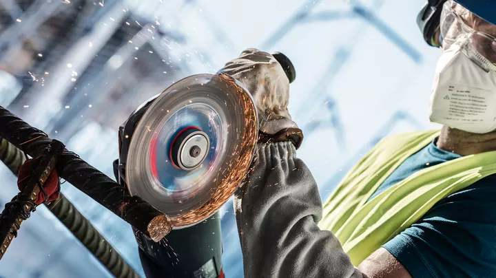 A person wearing safety equipment grinds metal rebar with sparks flying.