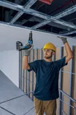 Person wearing safety equipment installs drywall using a cordless screwdriver.