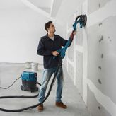 A person sands drywall with a drywall sander connected to a dust extractor.
