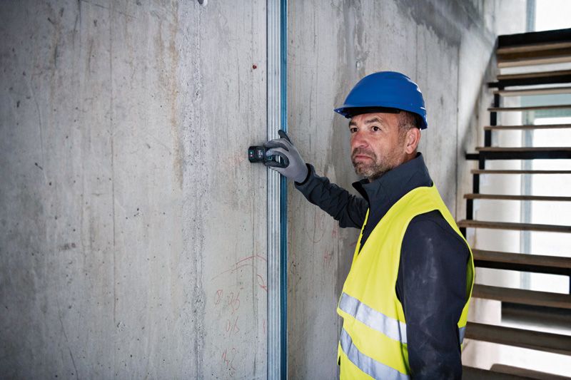 A person wearing safety equipment uses a laser measure against a concrete wall.