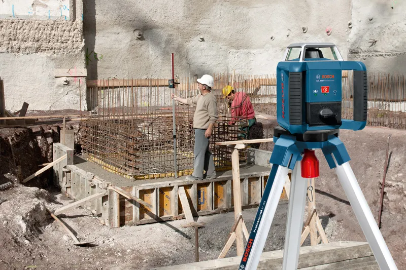 A person wearing safety equipment uses a laser leveling tool at a construction site.