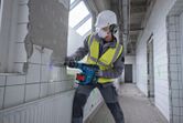 A person wearing safety equipment uses a cordless rotary hammer to break tiled wall.