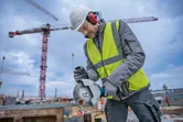 A person wearing safety equipment uses a cordless angle grinder to cut metal at a construction site.