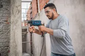 A person wearing safety equipment drills into a concrete wall with an impact drill.