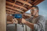A person wearing safety equipment drills into a wooden beam with an impact drill.
