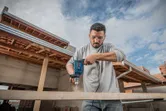 A person wearing safety equipment drills into a wooden beam at a construction site.