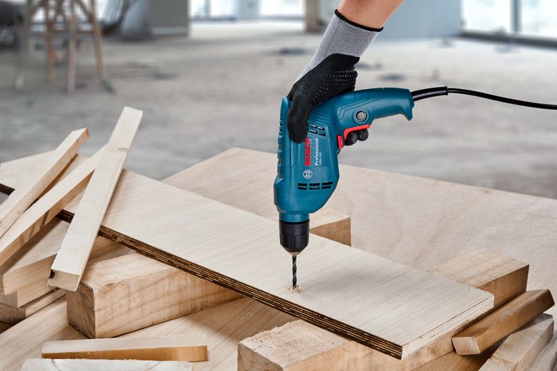 A person wearing safety equipment drills a hole in plywood on a workbench.