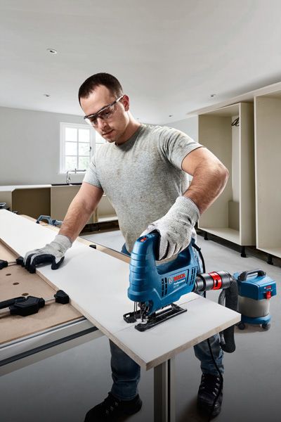 A person wearing safety equipment cuts a wooden board with a jigsaw in a workshop.