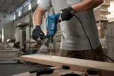 A person wearing safety equipment drills into a wooden board with an impact drill.