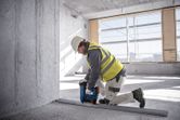 A person wearing safety equipment uses a cordless concrete nailer to fasten a metal track to the floor.