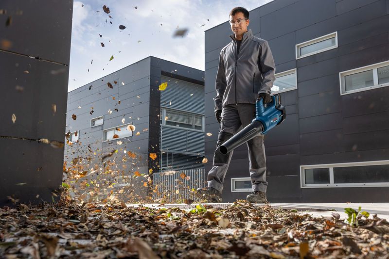 A person wearing safety equipment uses a cordless leaf blower to clear leaves near modern buildings.