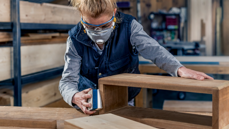 Person wearing safety equipment sands a wooden table in a workshop.