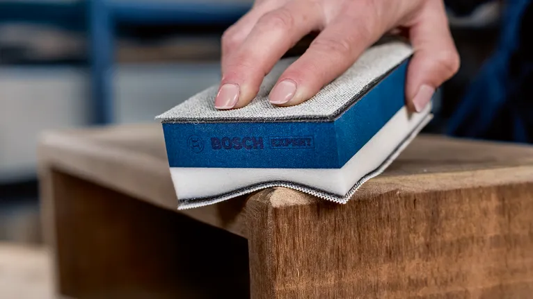 Person sanding a wooden surface with a sanding block.