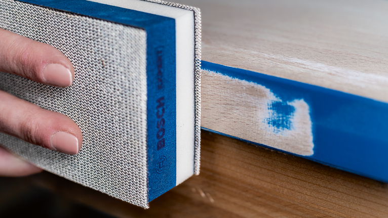 Person sanding a painted wooden edge with a sanding block.