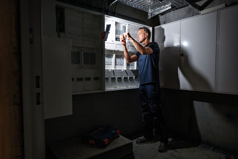 A person works on an electrical panel illuminated by a cordless work light.