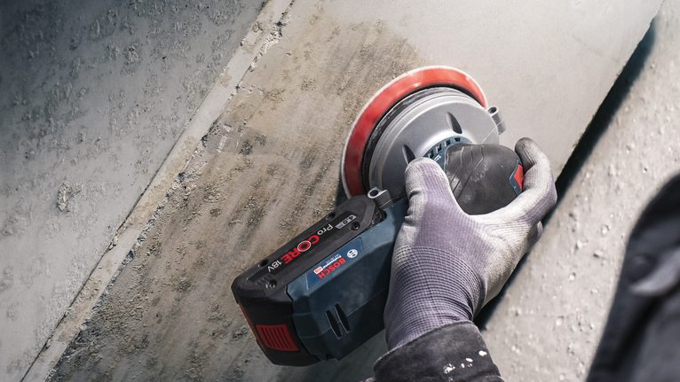 Person wearing safety equipment grinds a concrete wall with a cordless tool.