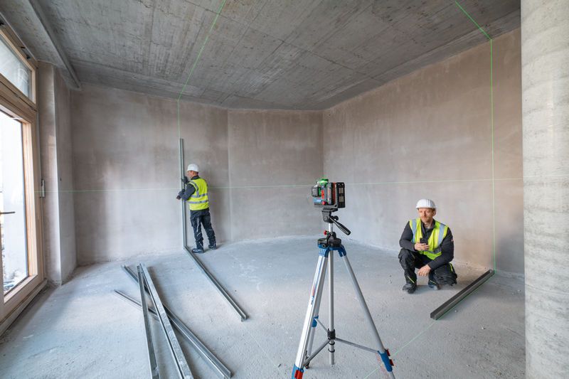 Two people wearing safety equipment use a laser leveling tool in an unfinished room.