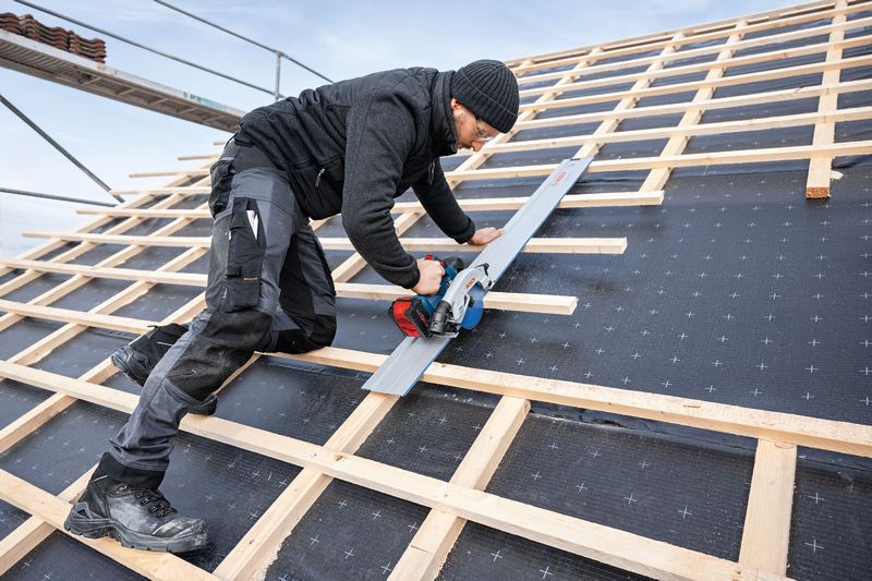 A person wearing safety equipment cuts wood on a steep roof using a cordless circular saw.