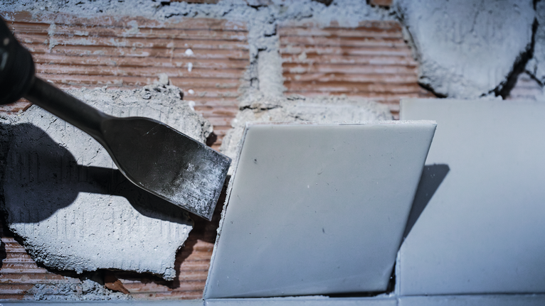 Chisel tool removing a wall tile, dust and debris visible on the surface.