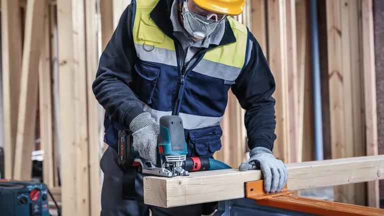 A person wearing safety equipment uses a jigsaw to cut a wooden beam.