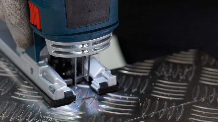 Person wearing safety equipment operates a jigsaw to cut a patterned metal sheet.