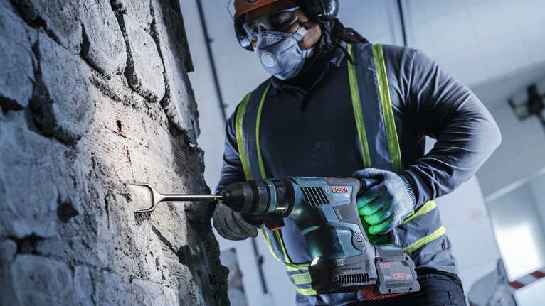 A person wearing safety equipment uses a cordless rotary hammer to chisel a concrete wall.