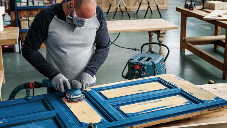 A worker wearing safety equipment sands an old wooden door in a workshop.