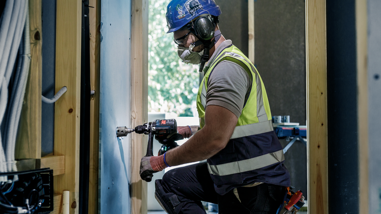 A person wearing safety equipment drills a hole in a wall at a construction site.