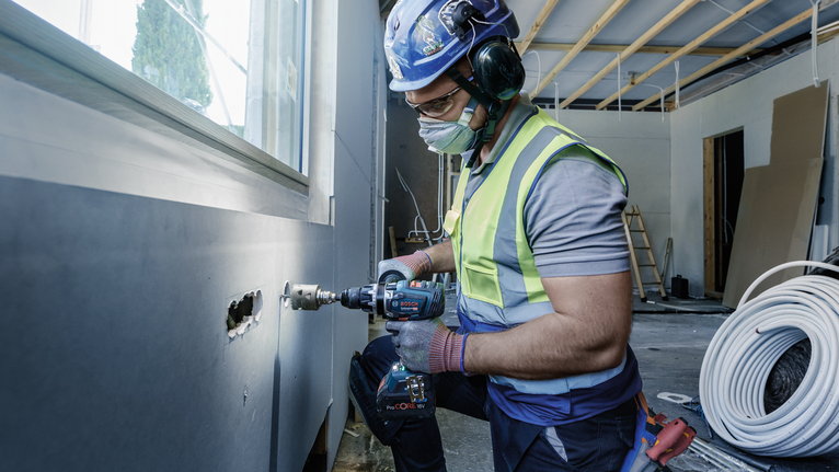 A person wearing safety equipment drills a hole in a wall at a construction site.