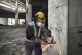 A person wearing safety equipment grinds rebar on a concrete wall with a cordless angle grinder.