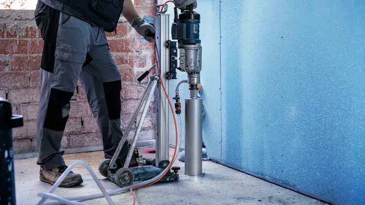 A person wearing safety equipment drills into concrete with a mounted coring machine.