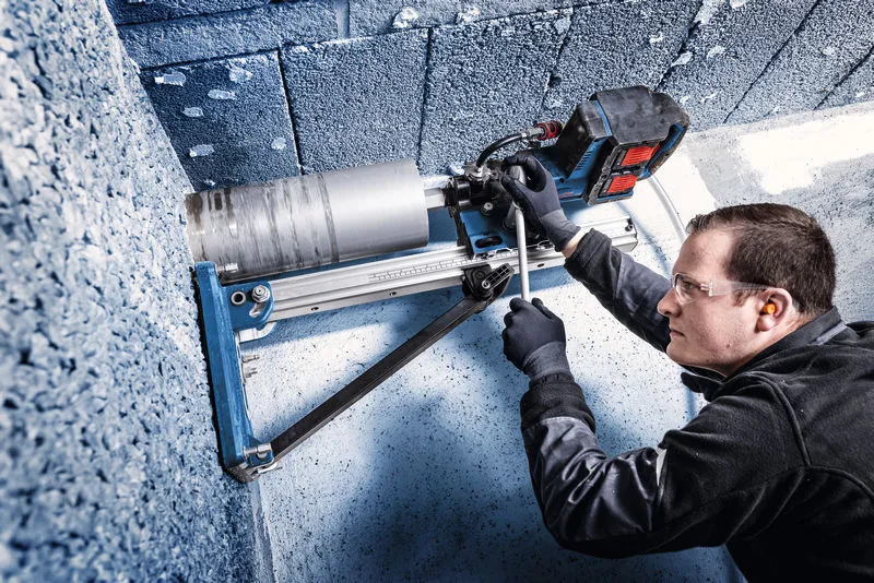 A person wearing safety equipment operates a mounted diamond drill on a concrete wall.